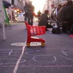 Man sieht einen Spielzeugeinkaufwagen auf Louisenstraße. Auf der autofreien Straße sitzen Menschen und essen.