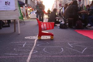 Man sieht einen Spielzeugeinkaufwagen auf Louisenstraße. Auf der autofreien Straße sitzen Menschen und essen.