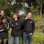 Fünf Menschen stehen in einem Garten. Alle schauen in unterschiedliche Richtungen, bloß nicht in die Kamera. Zwei davon rauchen.