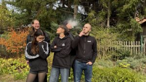 Fünf Menschen stehen in einem Garten. Alle schauen in unterschiedliche Richtungen, bloß nicht in die Kamera. Zwei davon rauchen.